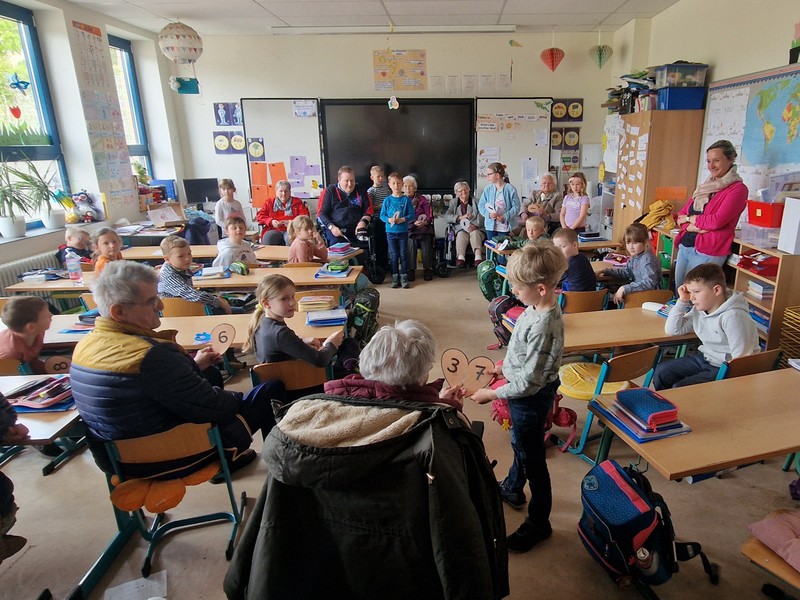 Bild zeigt Senior*innen und Grundschüler beim gemeinsamen lösen von Aufgaben in dem Klassenzimmer