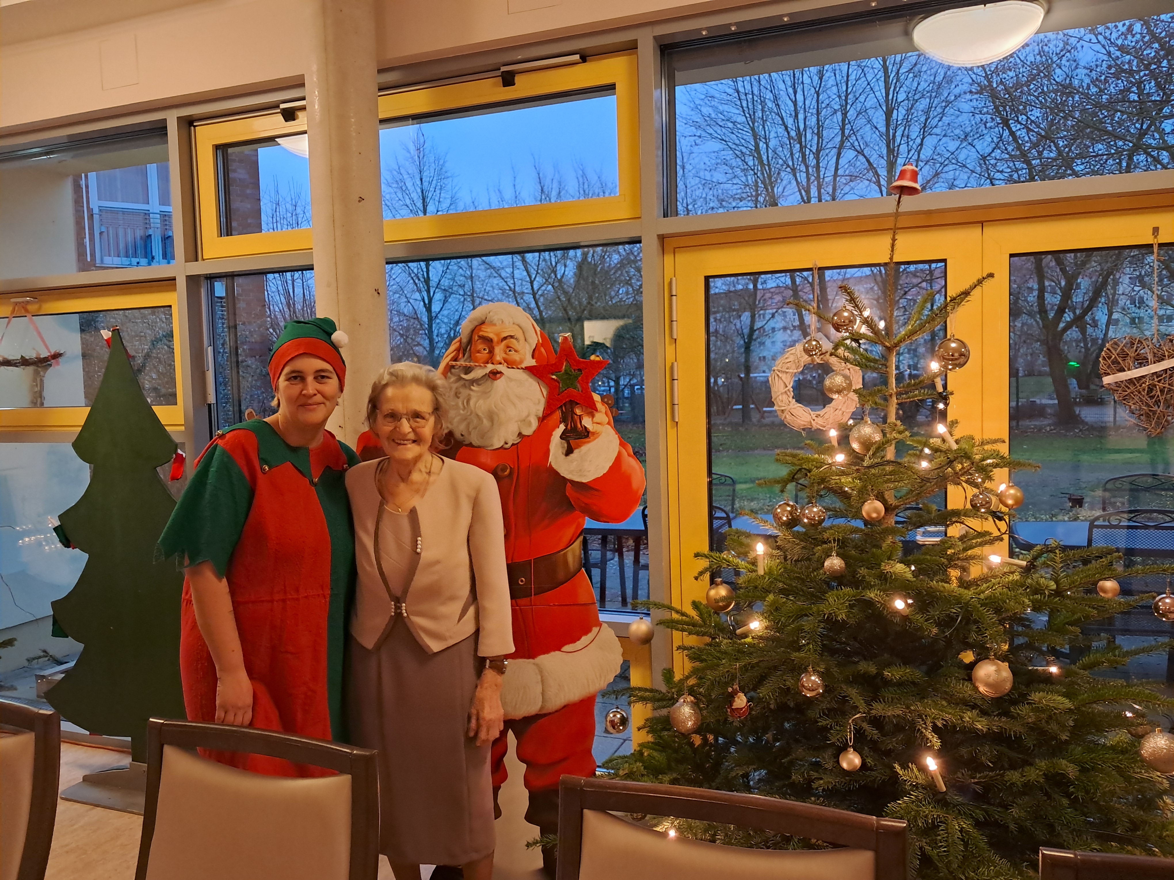 Bild zeigt Mitarbeiterin Nicole Reinholdt mit Tagesgast Christa Wähner und dem Weihnachtsmann vor dem Tannenbaum
