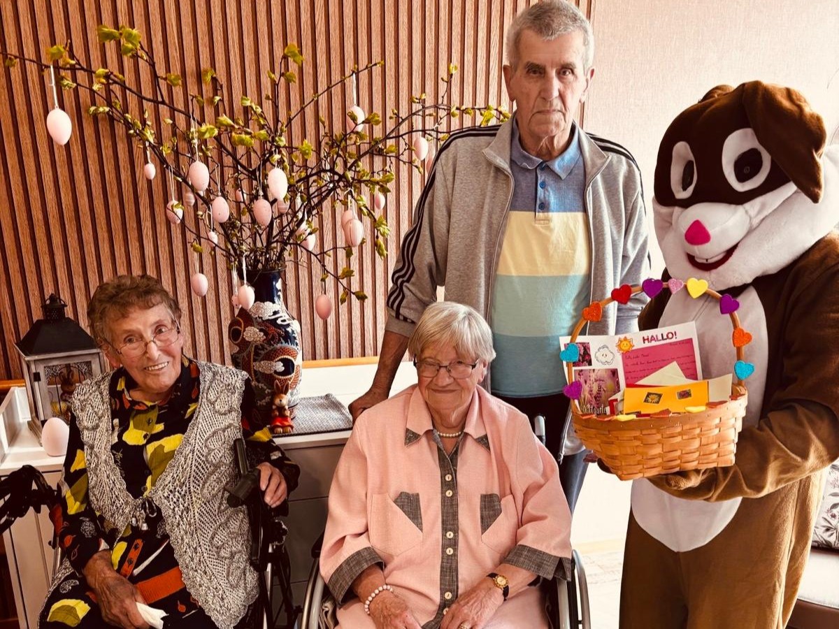 Bild zeigt 3 Bewohnende mit einem daneben stehenden "Osterhasen" (Mitarbeiter in einem Kostüm) vor einem bunt geschmückten Birkenstrauß. Der Osterhase hat ein Körbchen in den Pfoten, in dem die ganze Post liegt.